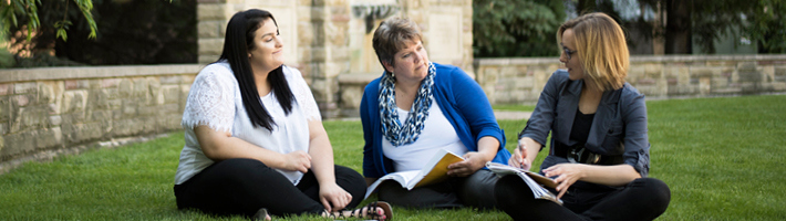 Three ladies sitting on the lawn in front of the a water fountain.  Two ladies have books open on their laps.