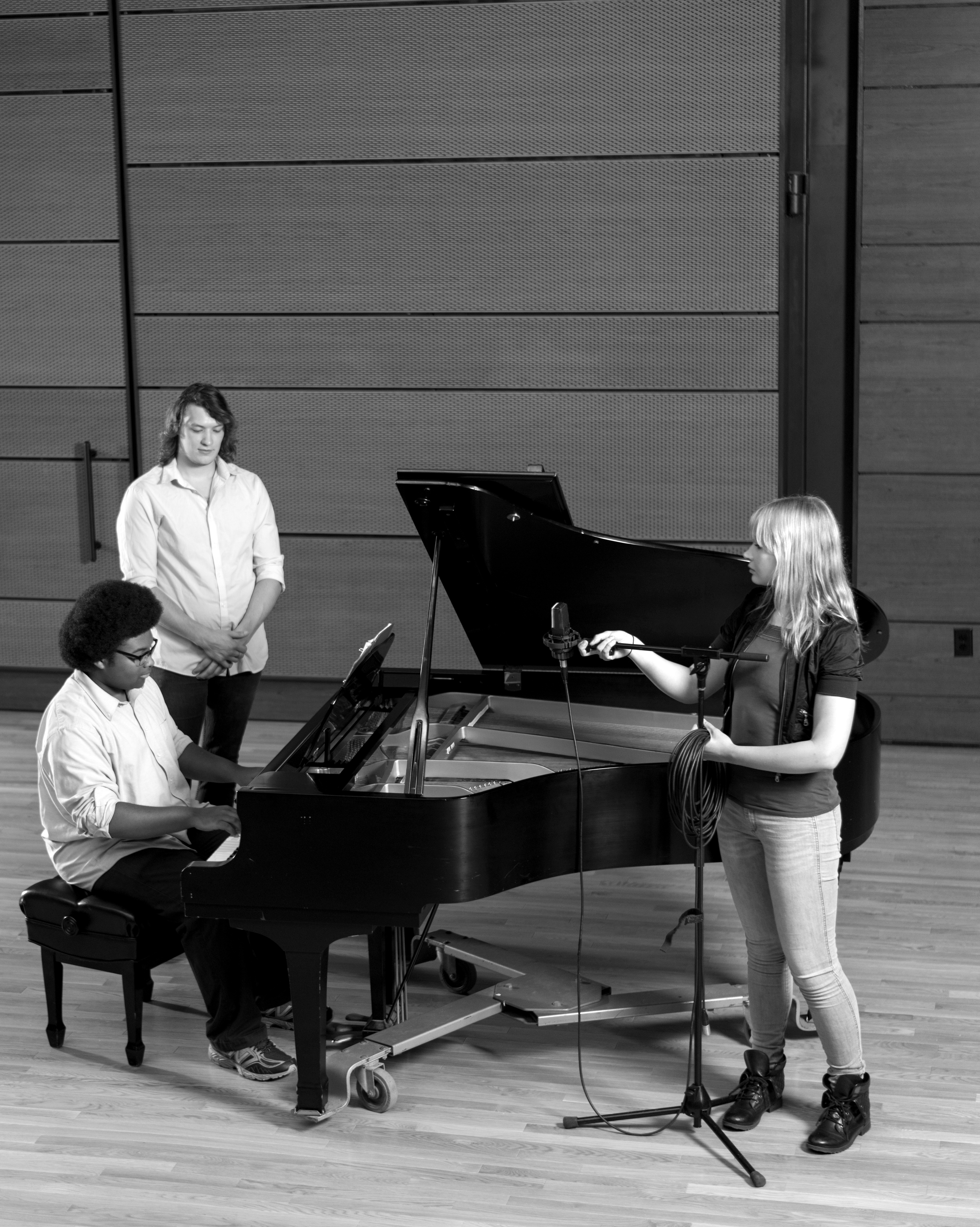 Three GRCC Music students in the recital hall.  One student is playing a piano, while one student is singing. 