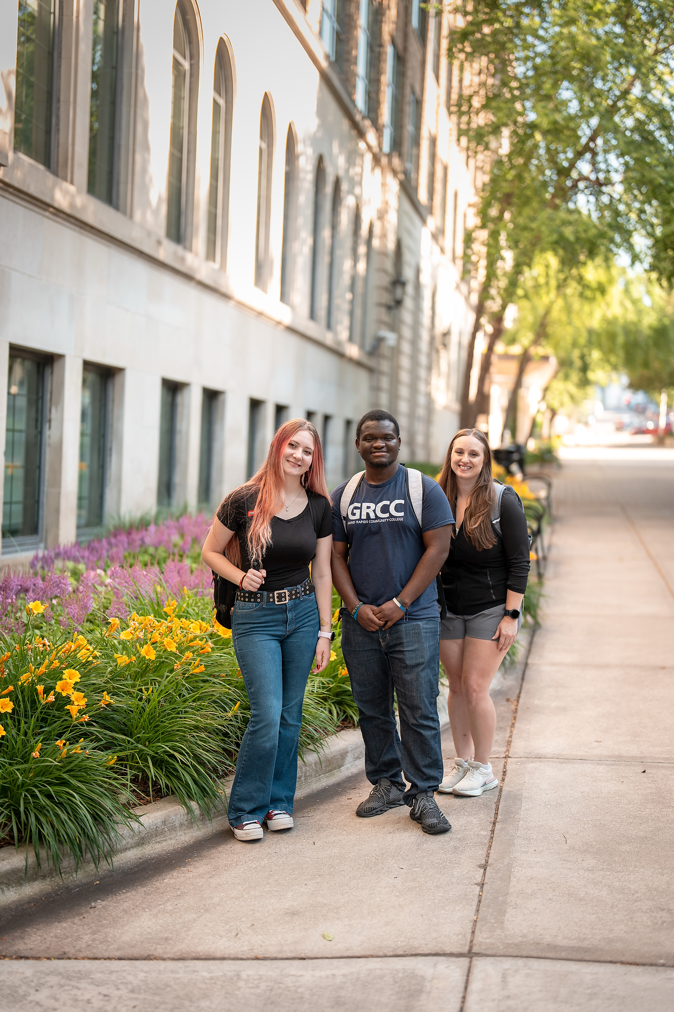 Three smiling students standing together on a sunny sidewalk at Grand Rapids Community College (GRCC). They are positioned in front of a stone campus building and a vibrant garden of yellow and purple flowers.