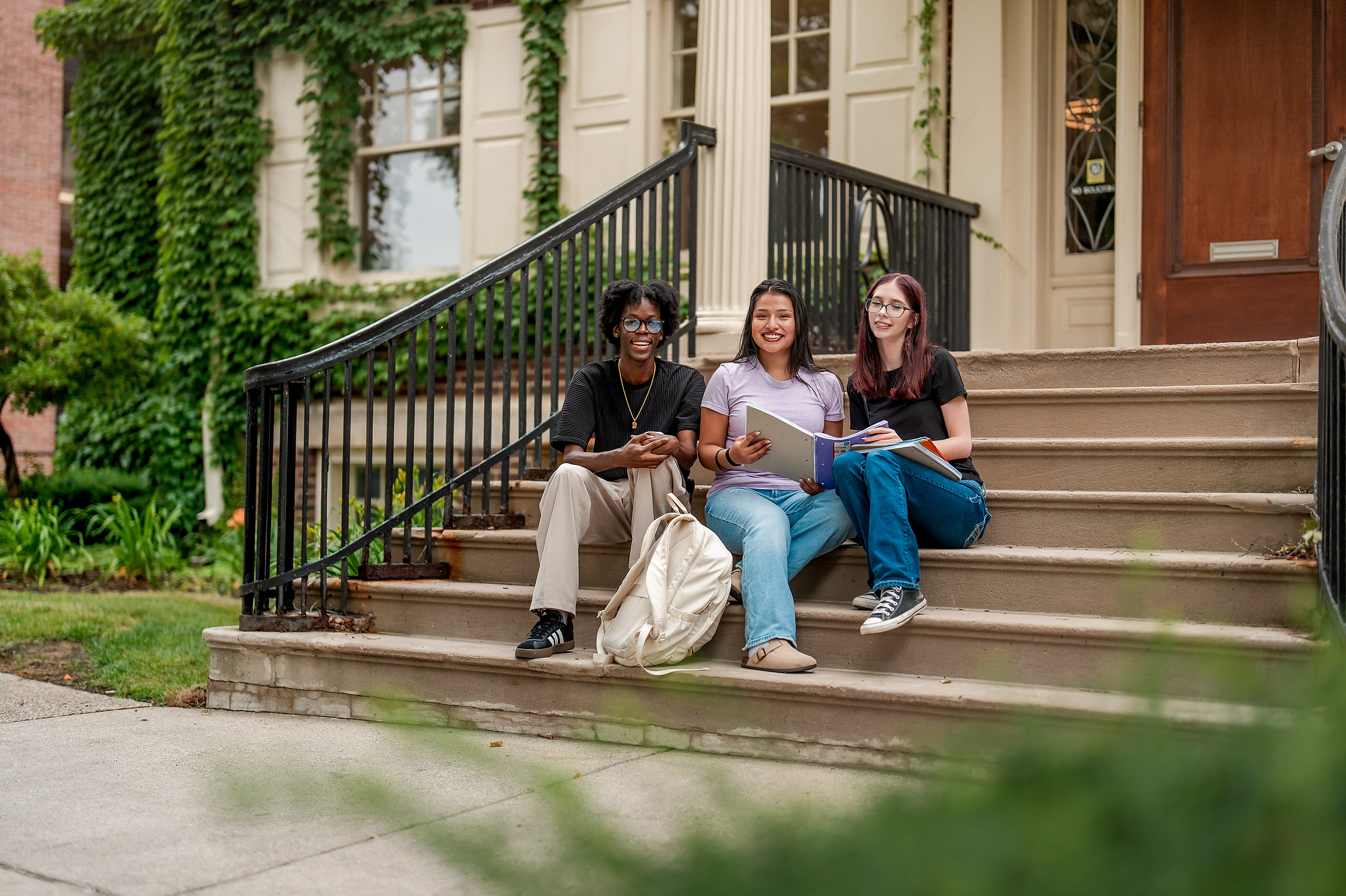 Three smiling students sitting on the concrete steps of a campus building at Grand Rapids Community College. Two of the students are looking at an open binder together while the third looks toward the camera.