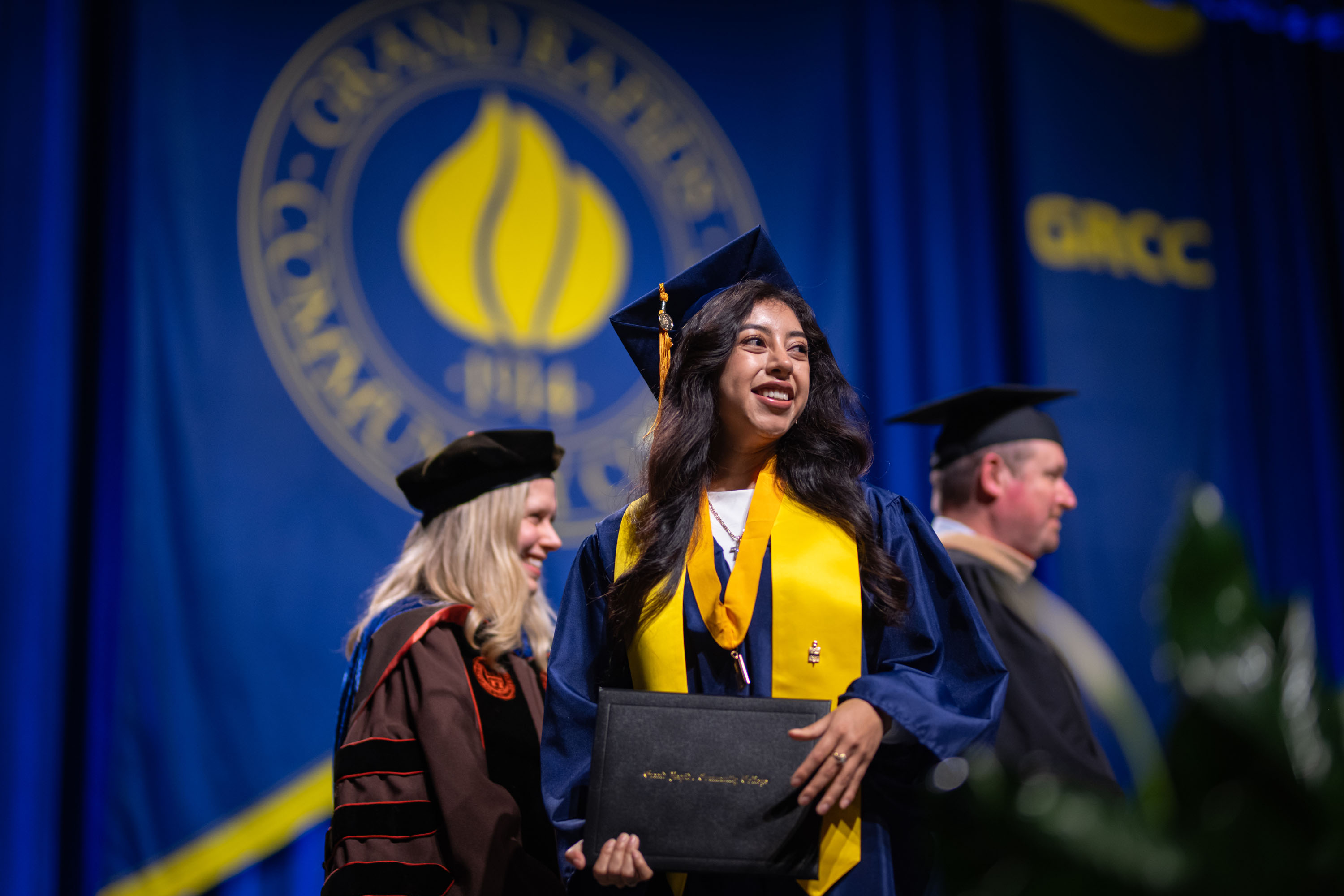 A smiling graduate in a navy blue cap and gown with a gold stole holds her diploma cover during a commencement ceremony. She stands on a stage in front of a large blue backdrop featuring the Grand Rapids Community College (GRCC) seal.