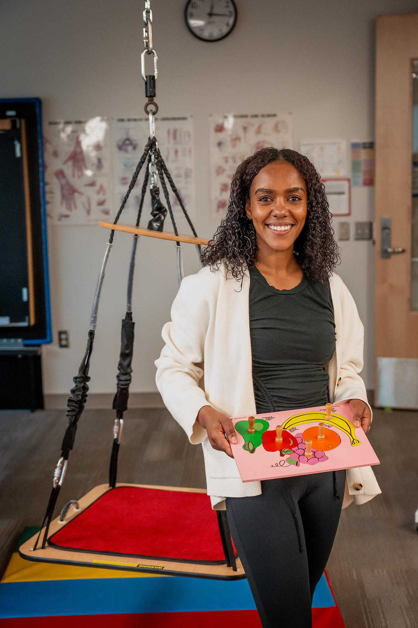 A smiling student in an Occupational Therapy Assistant (OTA) lab holds a wooden fruit puzzle. Behind her is a therapeutic platform swing and anatomical posters on the wall.