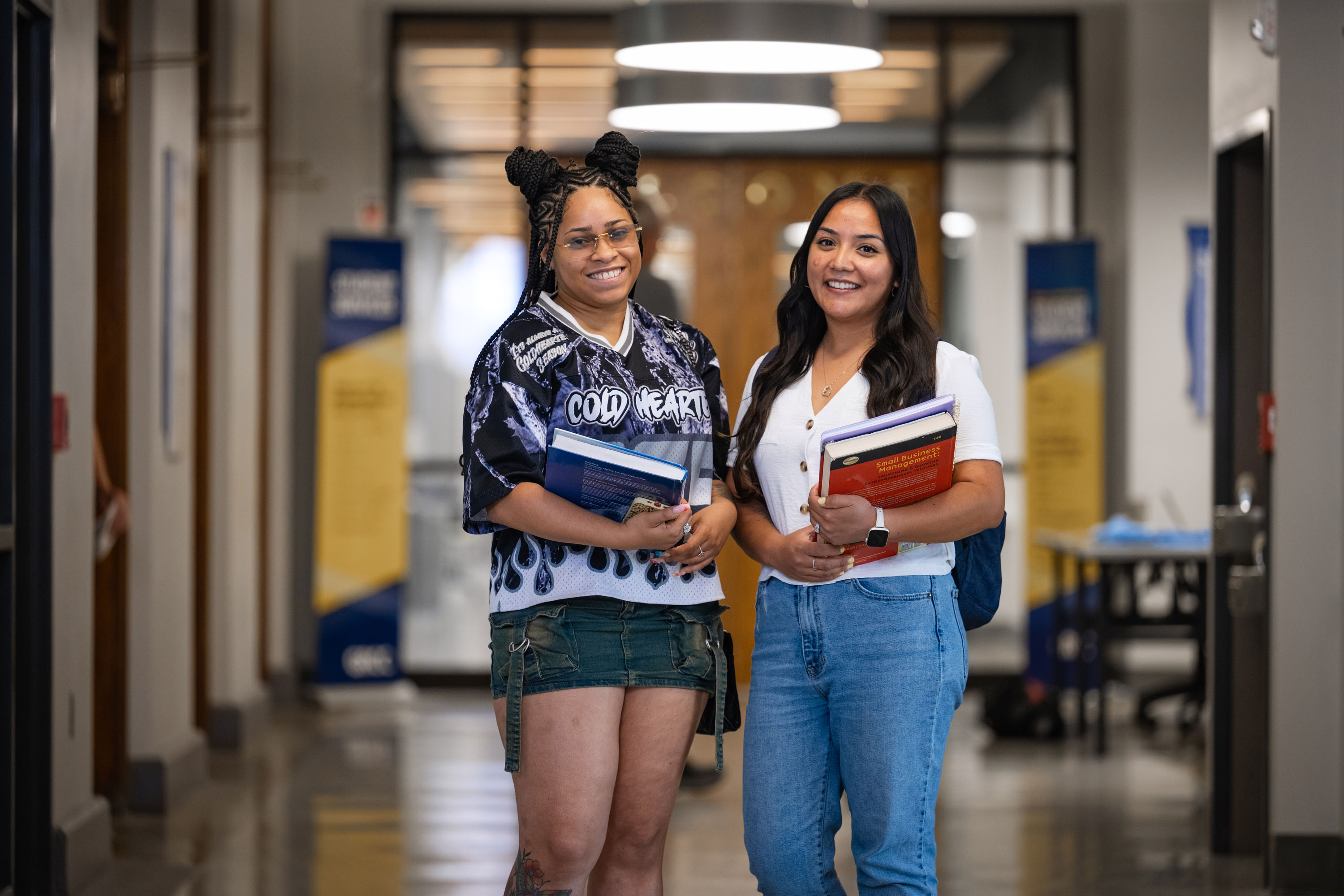 Two smiling students stand together in a brightly lit hallway at Grand Rapids Community College, both holding textbooks and ready for class.