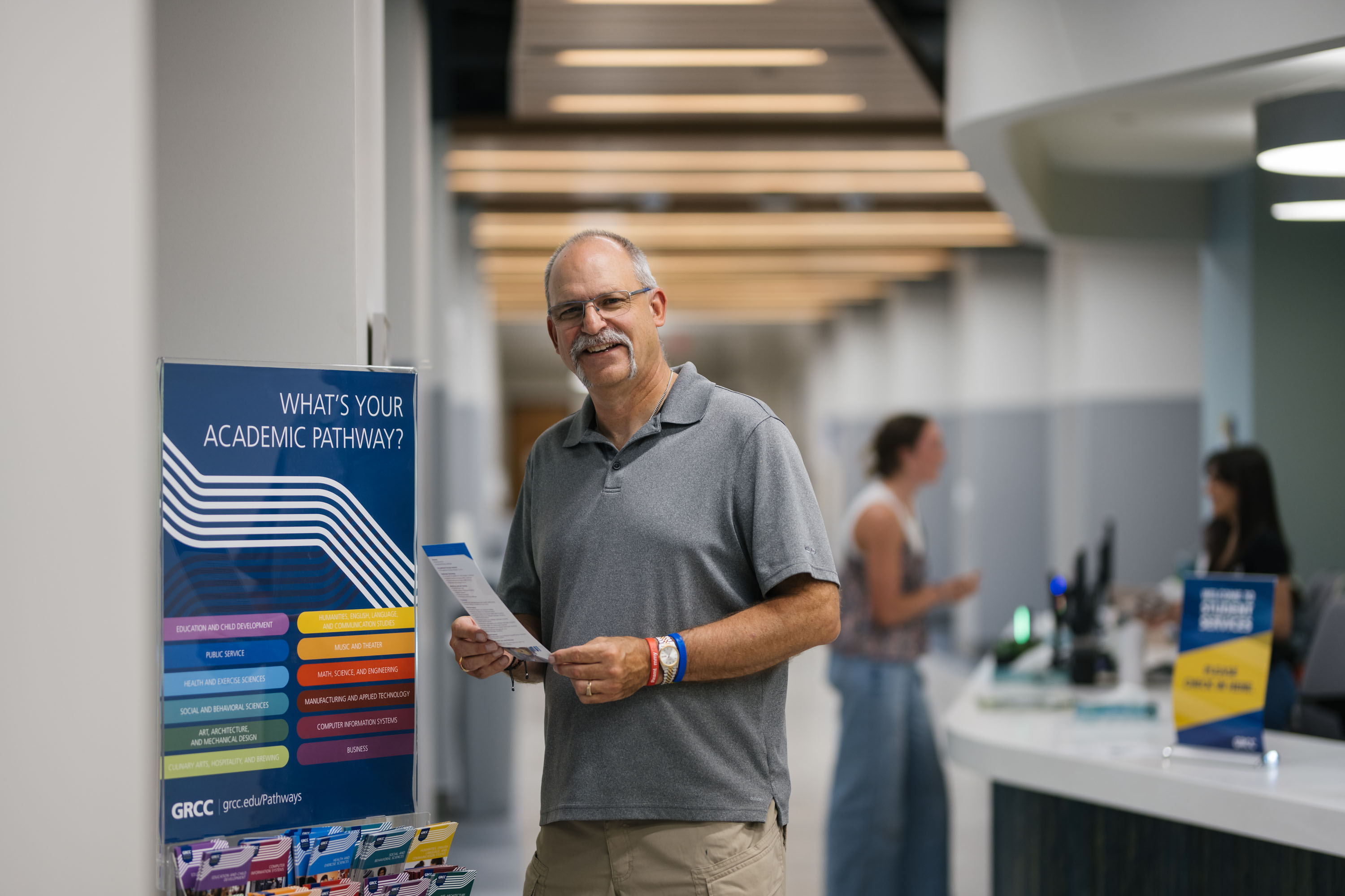 A smiling man stands in the Grand Rapids Community College (GRCC) Enrollment Center next to an "Academic Pathway" sign. He is holding an informational brochure, with a service desk and other staff visible in the blurred background.