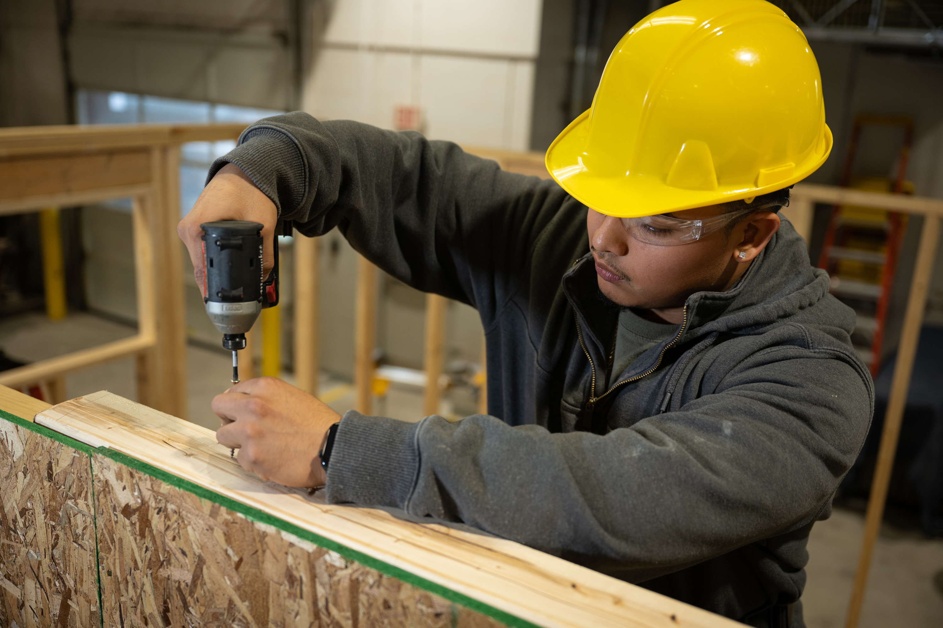 A student in a residential construction program wearing a yellow hard hat and safety glasses while using a power drill to drive a screw into a wooden wall frame.