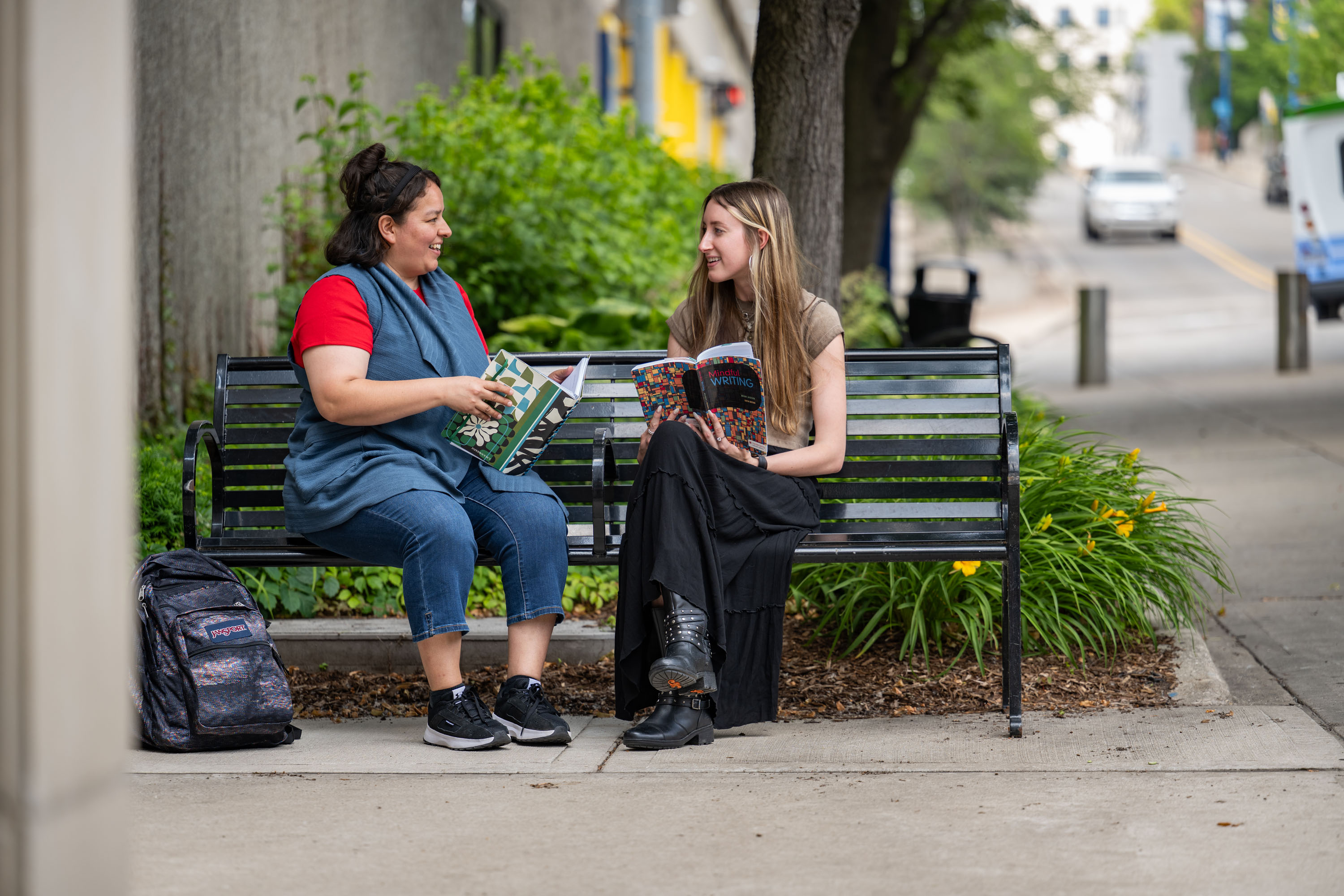  Two students sitting on a black metal park bench on a campus sidewalk, smiling and talking while holding open books. A backpack sits on the ground nearby.