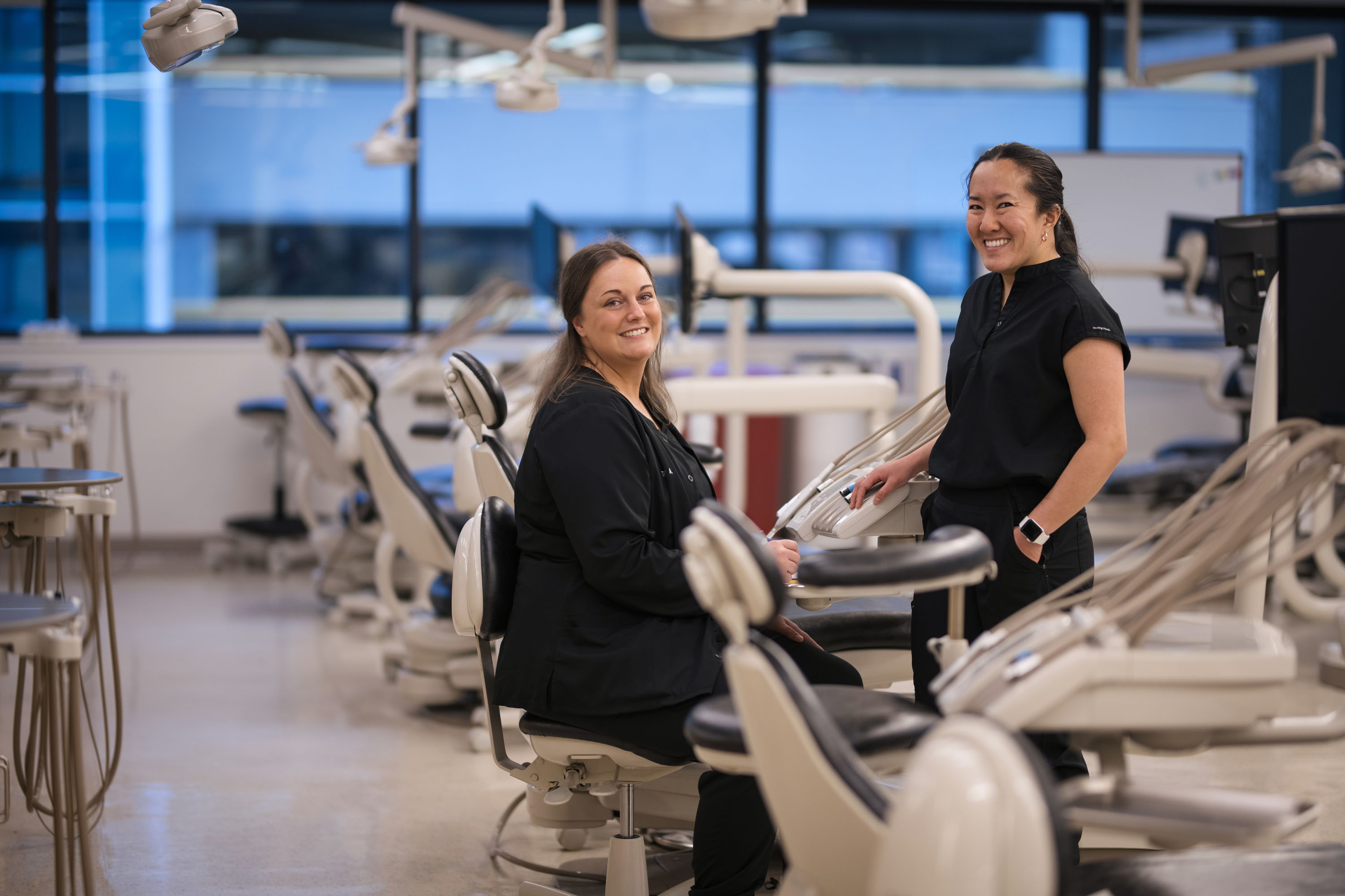 Two smiling dental hygiene students in black scrubs posing in a modern, multi-station campus dental clinic at Grand Rapids Community College.