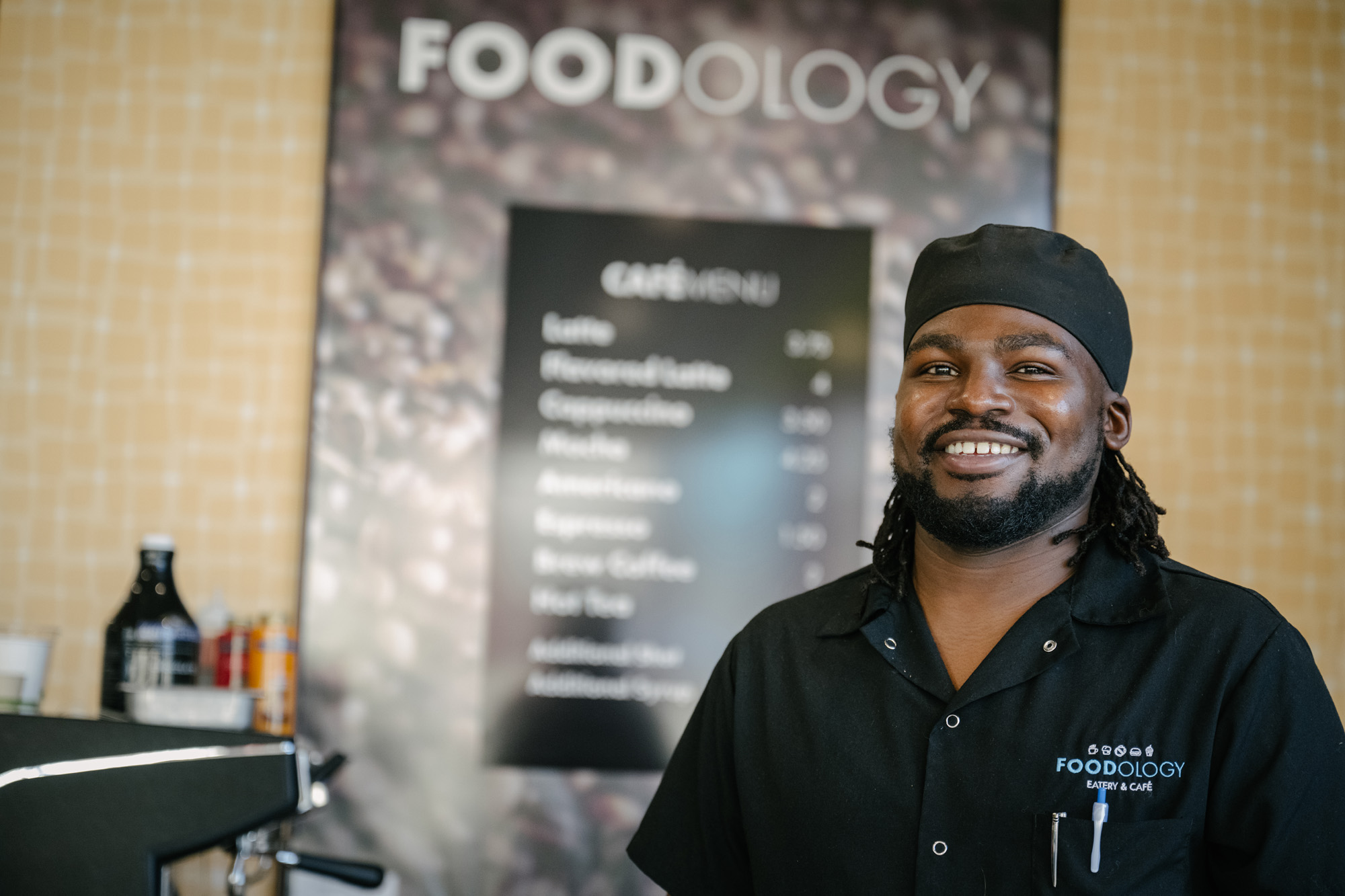 A smiling culinary student wearing a black chef's hat and a black uniform shirt with the "Foodology Eatery & Café" logo. He stands in front of a café menu listing various coffee drinks.