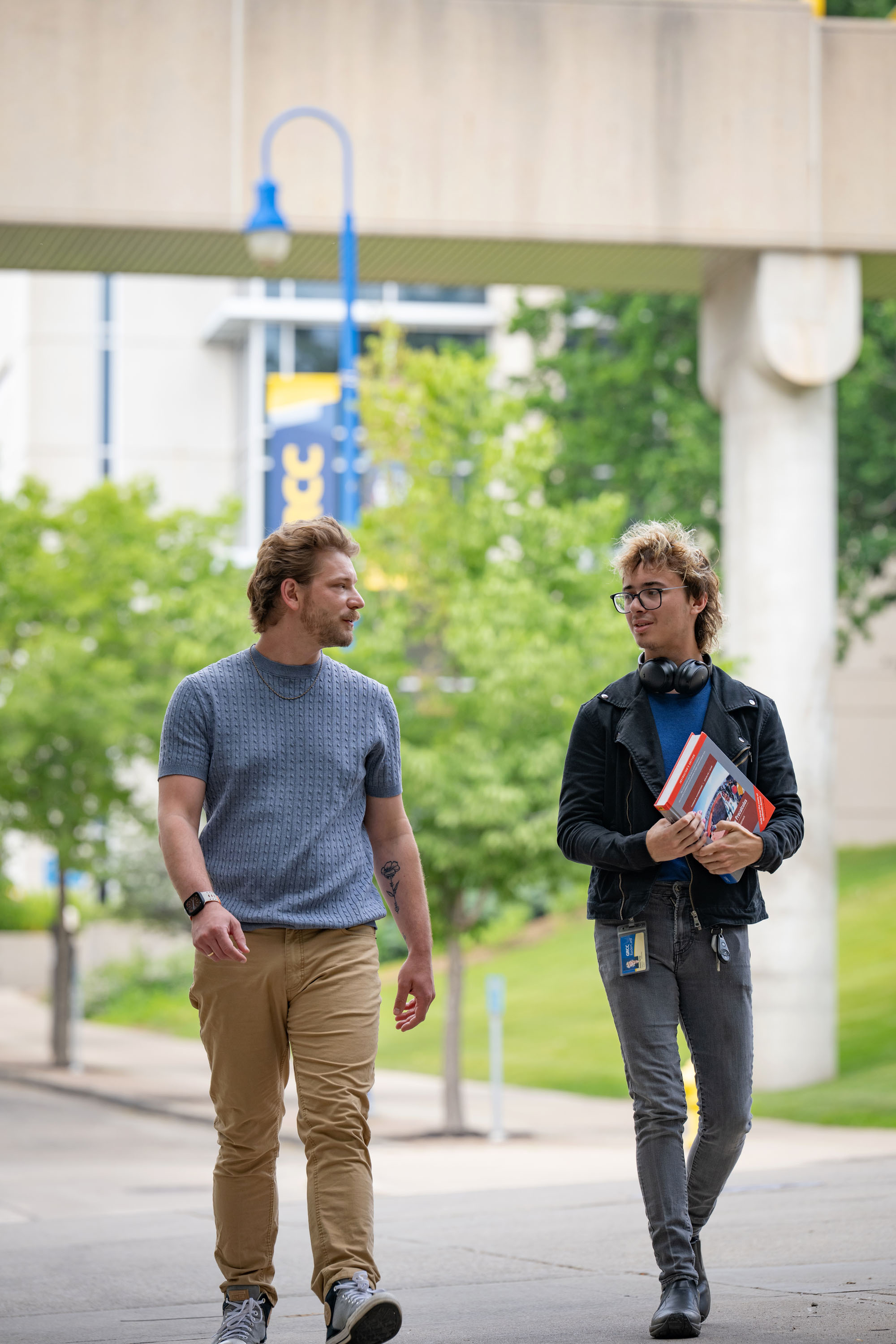 Two students walking and talking on a campus sidewalk at Grand Rapids Community College, one holding a textbook. A skywalk and green trees are visible in the background.