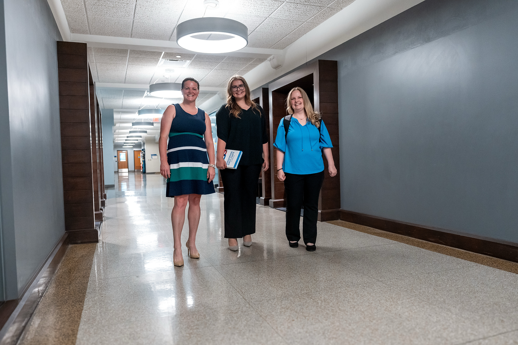 Three women standing and smiling in a long, brightly lit hallway at Grand Rapids Community College. One student in the center is holding a textbook.