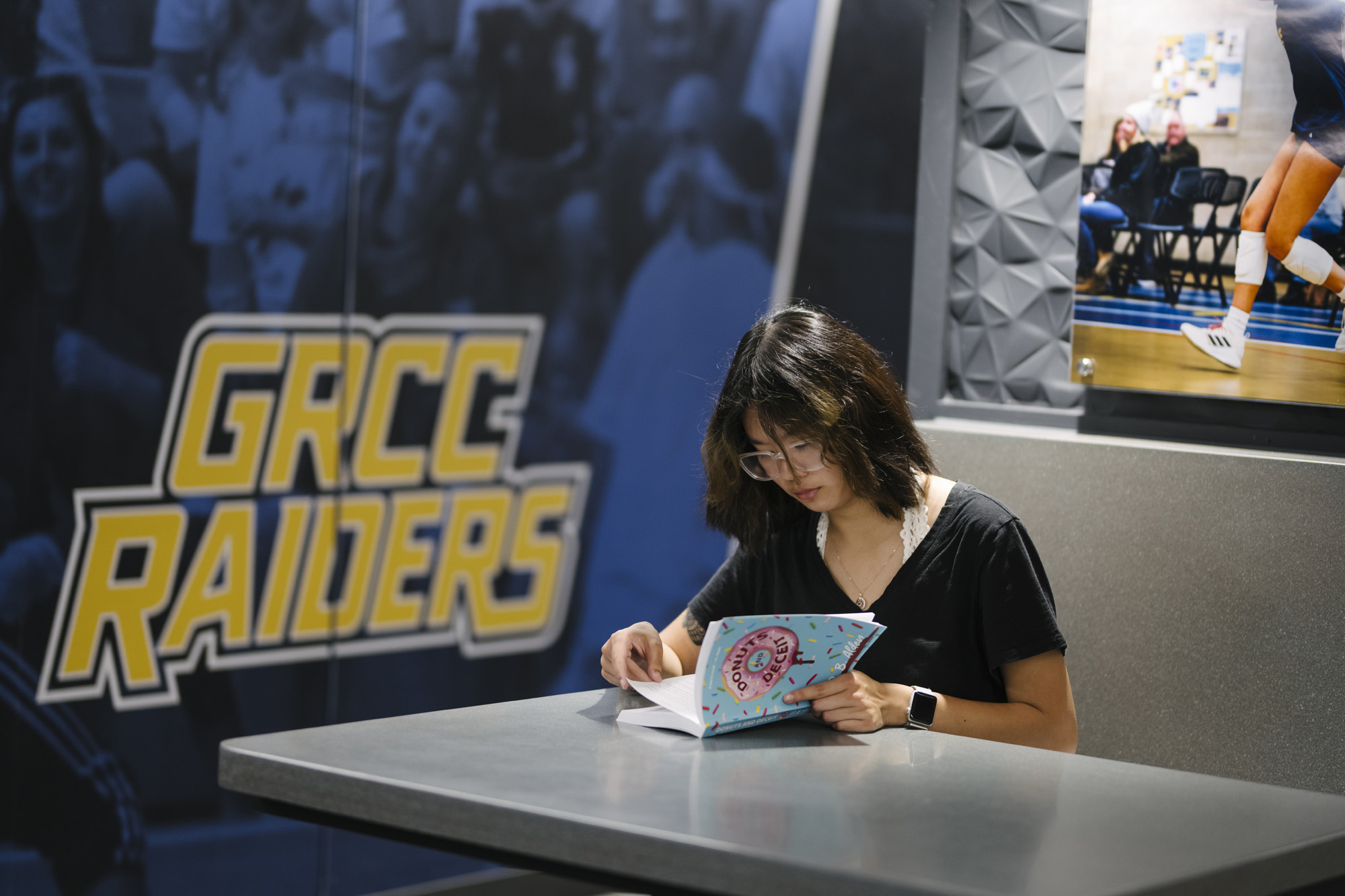 A student sitting at a grey table in a campus lounge, focused on reading a colorful book titled "Donuts and Deceit." A large "GRCC Raiders" logo is visible on the wall behind her.