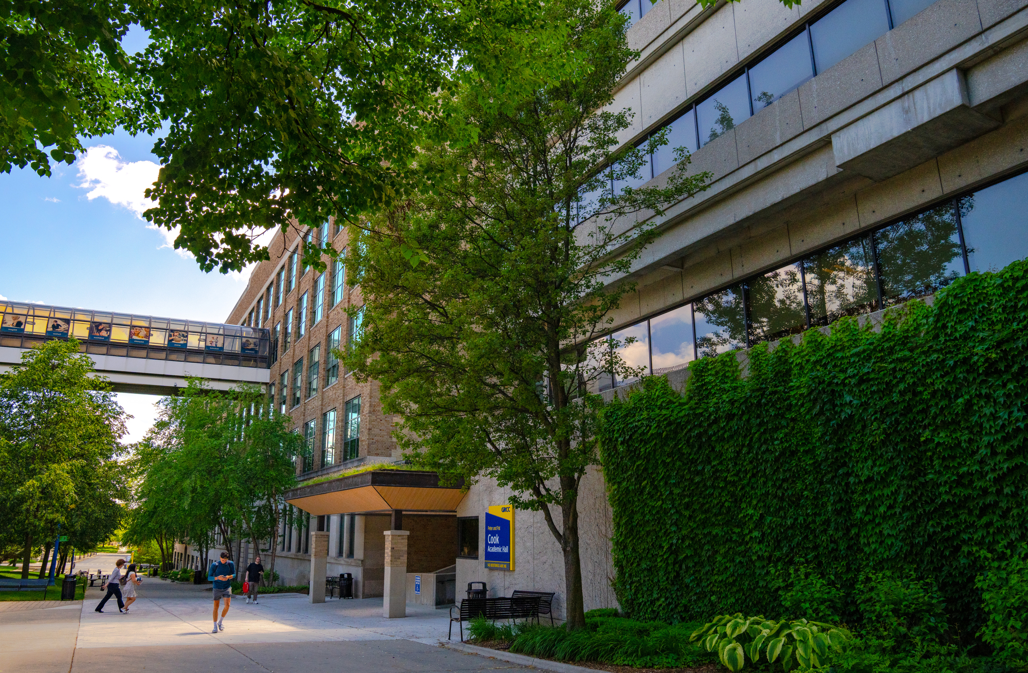 A view of the Cook Academic Hall at GRCC on a sunny day. The scene features a multi-story building with a skywalk, lush green trees, and an ivy-covered wall bordering a concrete campus walkway where students are walking.
