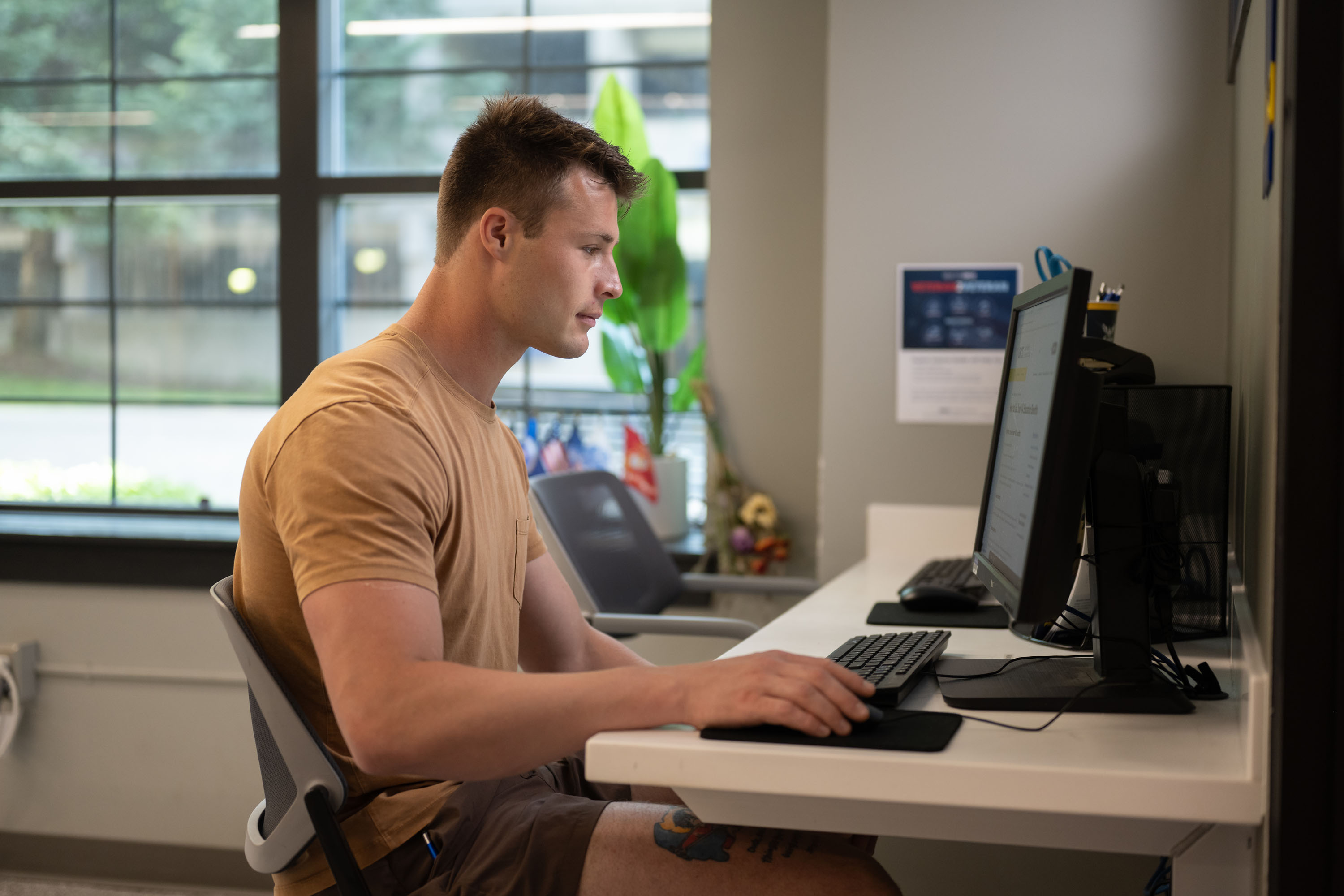 A male GRCC student sits and uses a desktop computer at a workstation in a modern office or computer lab, situated near a large window.